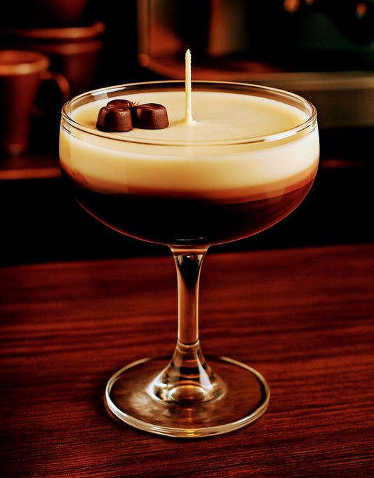 A large luxury candle styled like a layered espresso cocktail, featuring a creamy top layer with two chocolate pieces and a central wick, set in an elegant glass on a wooden surface. The candle evokes rich coffee and vanilla tones, blending visual sophistication with aromatic warmth.
