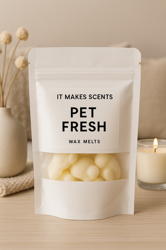 White resealable pouch of "Pet Fresh" wax melts from It Makes Scents, displayed on a light wood surface with cozy home accents including a lit candle and dried flowers. Label features bold black text on a white background, with wax melts visible through a clear window.
